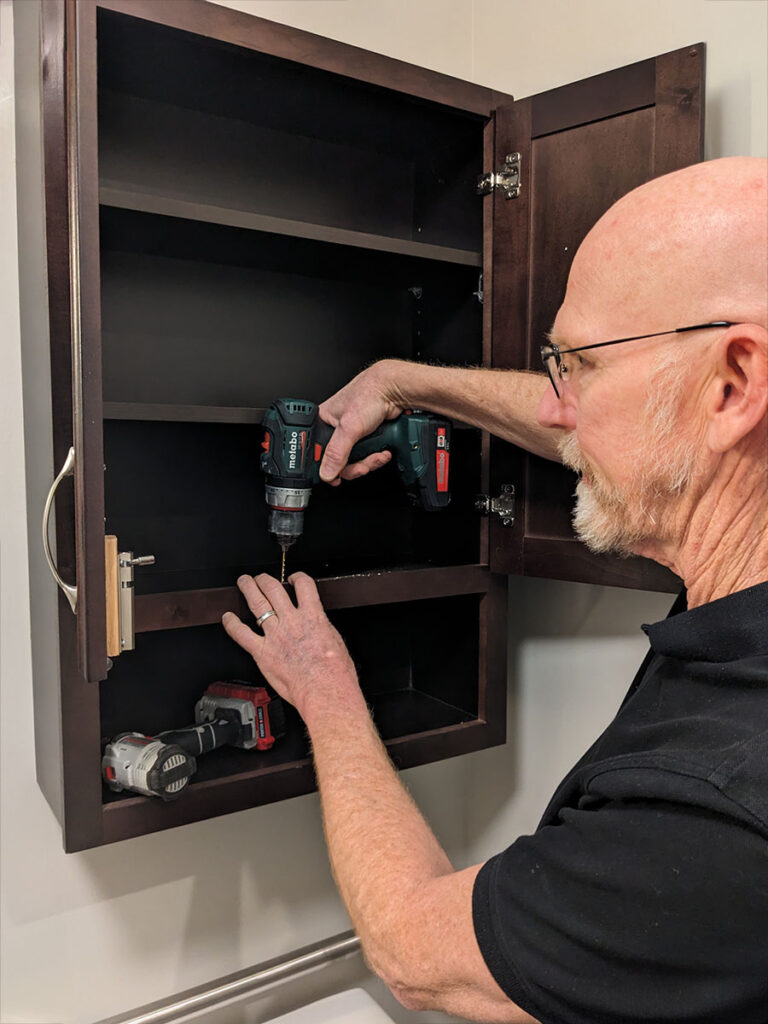 A man using a power tool on a dark brown wall cabinet.