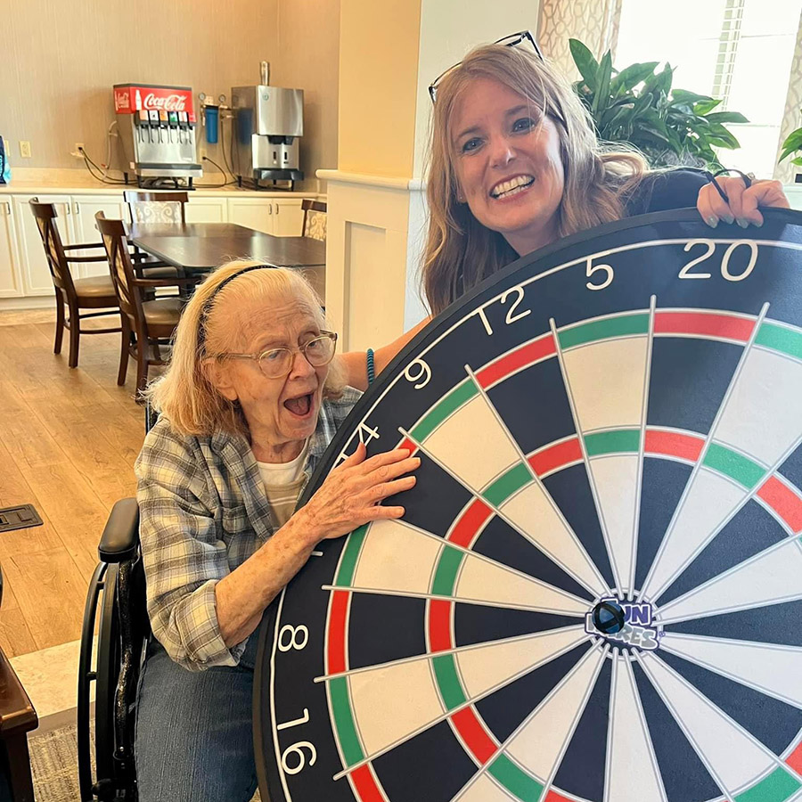 A woman in a wheelchair smiling, holding a dart board. Memory Care residents are having fun playing darts; she even made a bullseye!