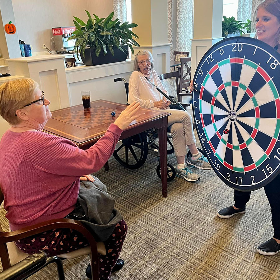 Woman holds dart board as two others sit in chairs, Memory Care residents enjoy game of darts.