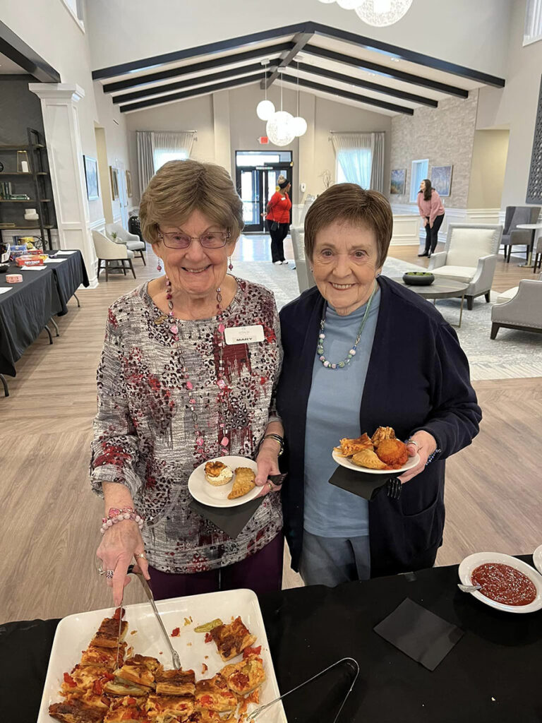 A delightful moment is captured as two elderly ladies are at a gathering at the senior community, dining to relish a scrumptious buffet, plates filled with mouthwatering food.