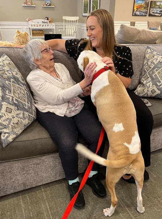 Jessica, the memory care director, sitting on a couch with a senior resident, both smiling and interacting with a large dog standing on its hind legs.