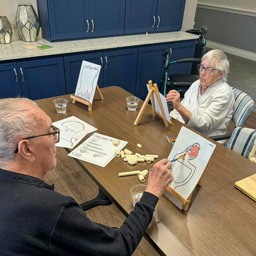 Two senior residents engaged in a painting activity, seated at a table with easels and art supplies, working on their artwork.