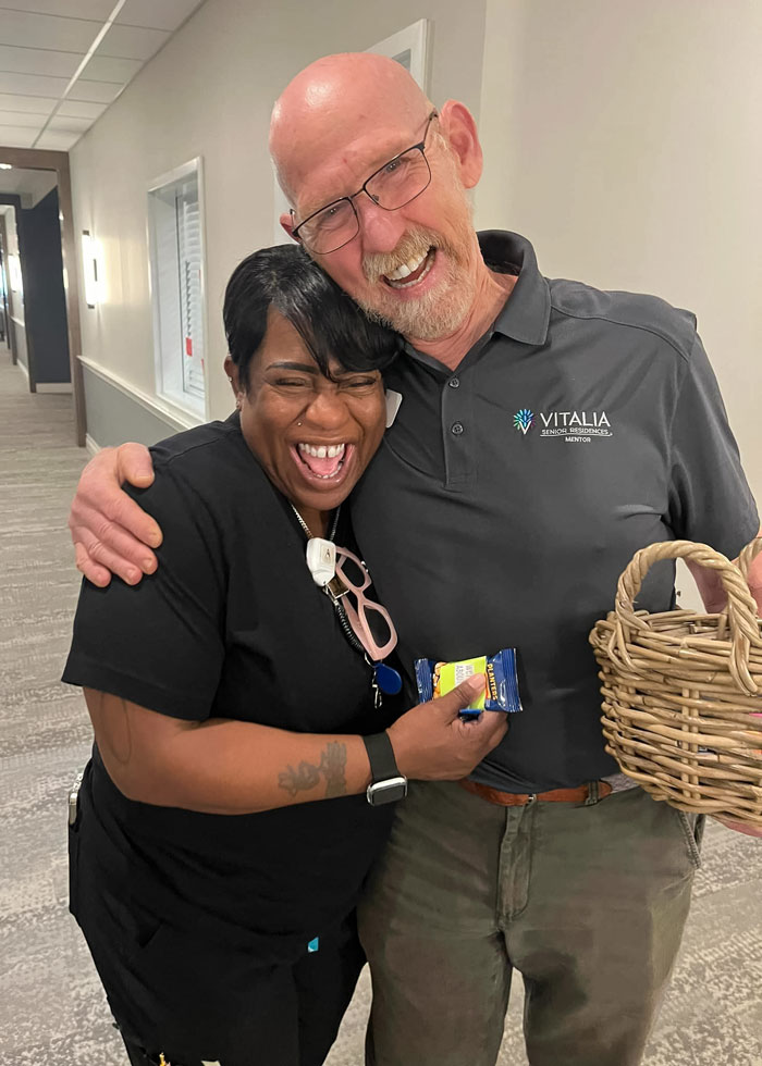 A joyful moment between two Vitalia Mentor team members, with a man in a Vitalia polo shirt hugging and smiling with a woman holding a snack bar and a small wicker basket.