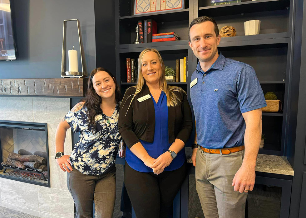 Three Vitalia Senior Living employees standing together in front of a bookshelf and fireplace, all wearing blue for Pride Week, showing solidarity and support.