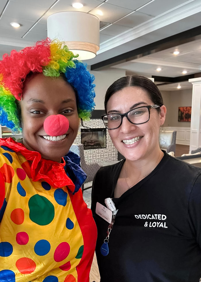 Two smiling women, one dressed as a colorful clown with a rainbow wig and red nose, and the other in a black shirt that reads 'DEDICATED & LOYAL' during Clown Week.