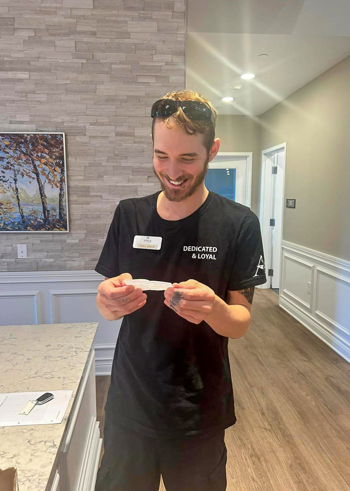 A Vitalia Mentor team member smiling while reading a note, wearing a 'Dedicated & Loyal' shirt, standing in a warmly lit room with a stone accent wall.