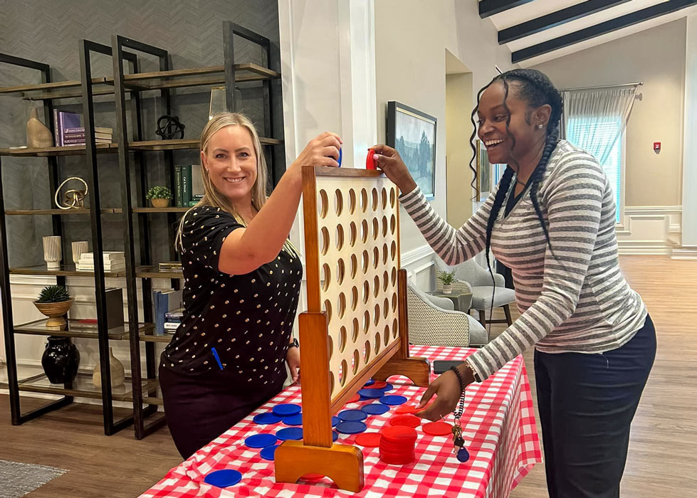 Two Vitalia Mentor team members happily playing a large Connect Four game during a summer cookout event, with a red and white checkered tablecloth on the table.