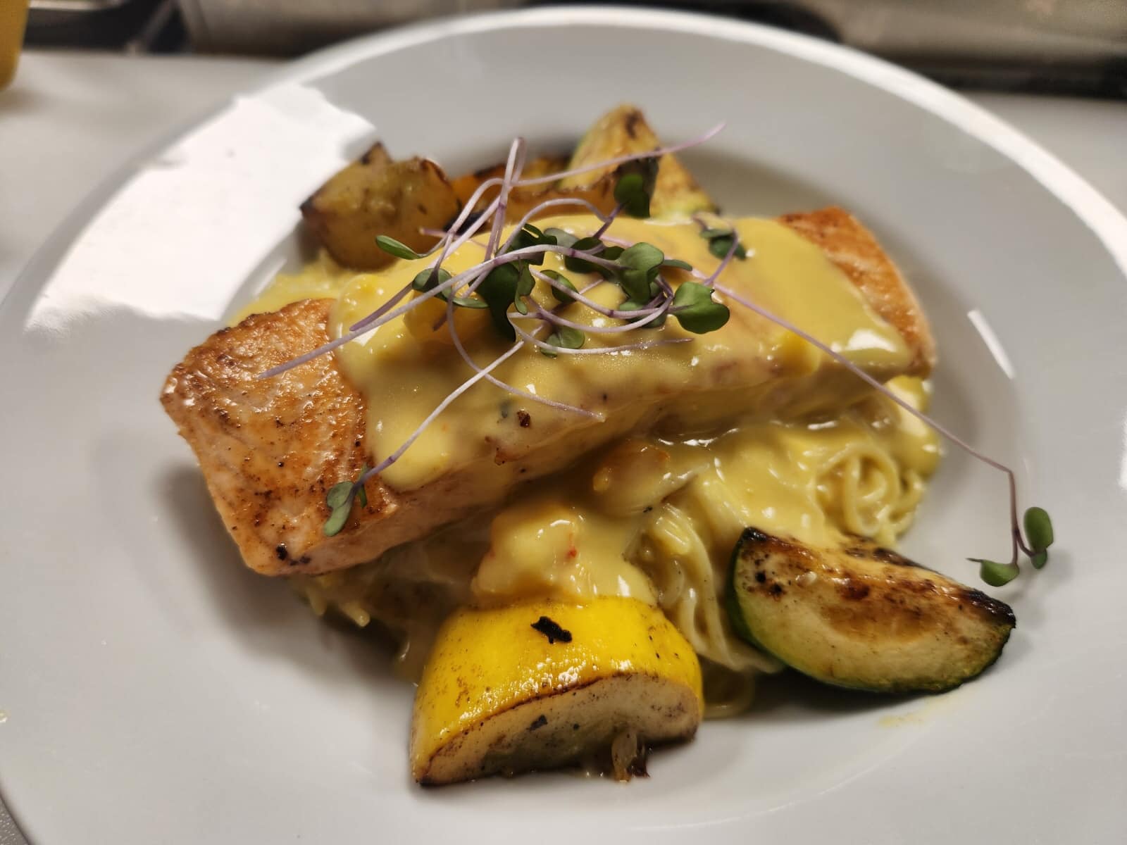 Grilled salmon topped with a creamy sauce and microgreens, served with roasted lemon and zucchini on a bed of pasta, offering a beautifully plated gourmet meal.