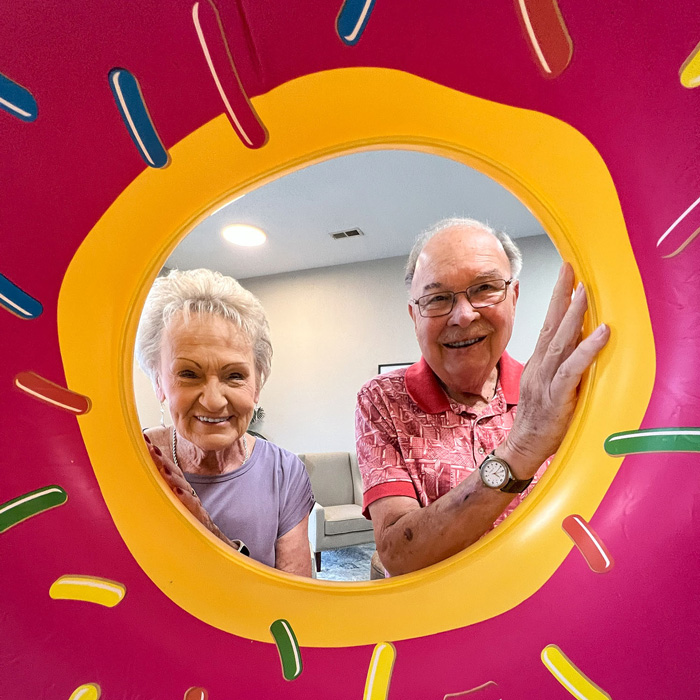 Two seniors smiling through a colorful donut-shaped pool float, capturing a fun and playful moment indoors, highlighting the joy and vibrancy of senior community living.