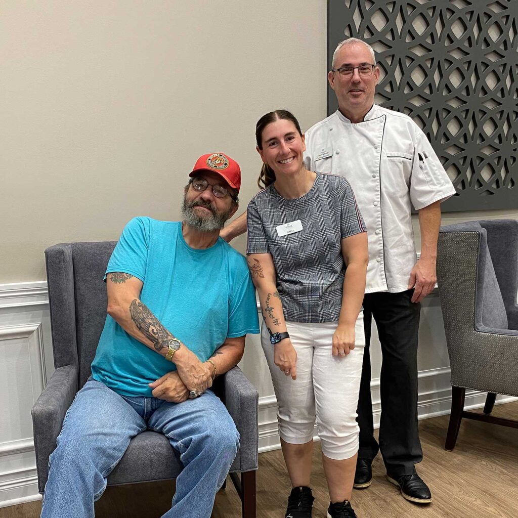 A senior resident in a red hat poses with two smiling team members, including a chef in a white coat, creating a warm moment of connection in the community.
