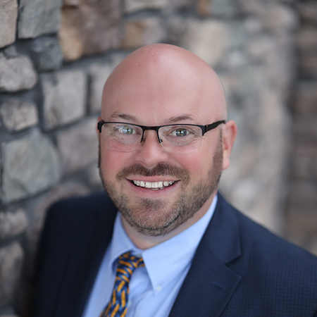 Mark Stapleton, Arrow Regional Director of Operations and acting Executive Director at Vitalia Mentor, smiling in a professional headshot. He is wearing glasses, a navy suit, a light blue shirt, and a patterned yellow and blue tie, standing against a stone wall background.