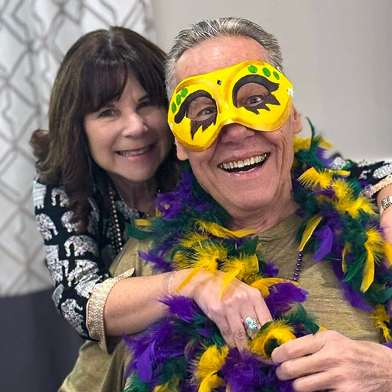 A man and woman smile brightly, the man dressed with mask and feather boa for Mardi Gras.