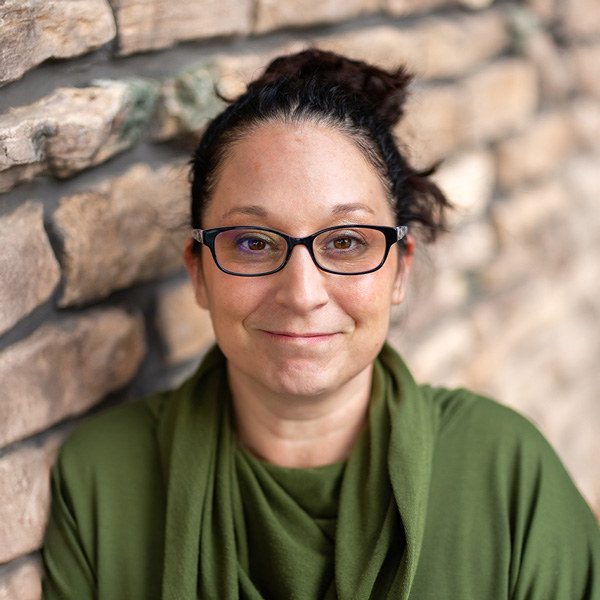 Andrea Zorik, Memory Care Director at Vitalia Mentor, smiling softly while wearing glasses and a green top, standing beside a textured stone wall.