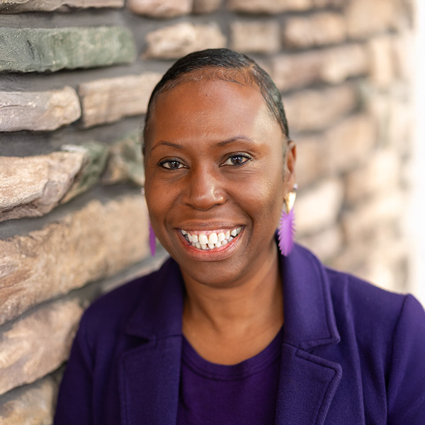 Carmella Pinesett, Resident Services Director at Vitalia Mentor, smiling in a purple top and blazer with matching earrings, standing beside a textured stone wall.