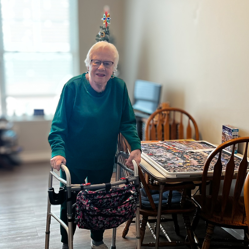 A senior woman smiles, standing next to a table topped with a large puzzle.