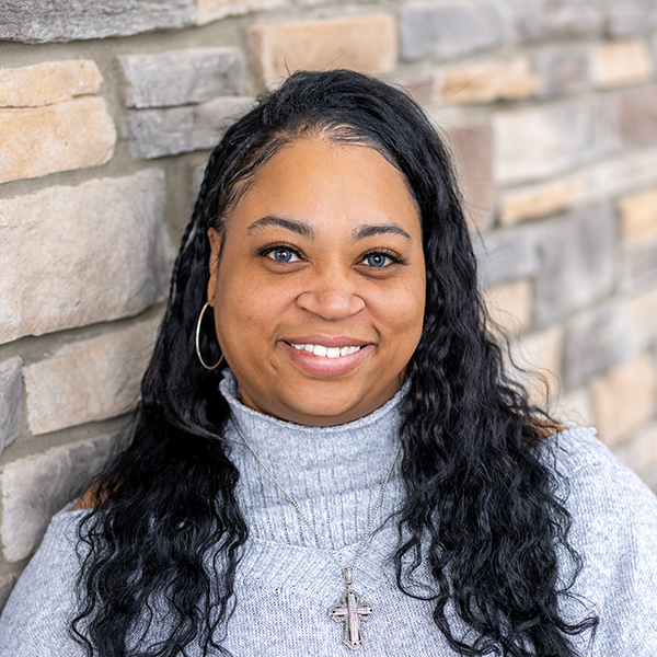 Tatianna Cotten, the Assistant Executive Director at Vitalia Mentor, smiles in a gray sweater while standing in front of a stone wall background.