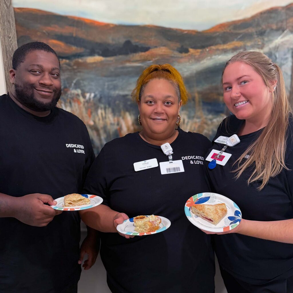 Three team members at Vitalia Mentor smile, holding plates of desserts during a team event.