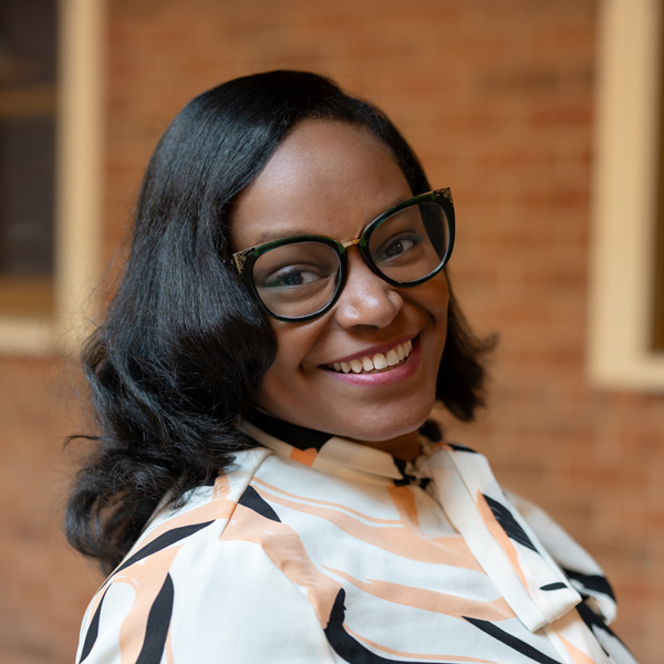 Corianne Lee, Business Office Director at Vitalia Mentor, smiling in a professional headshot, wearing glasses and a patterned blouse, with a softly blurred brick and window background.