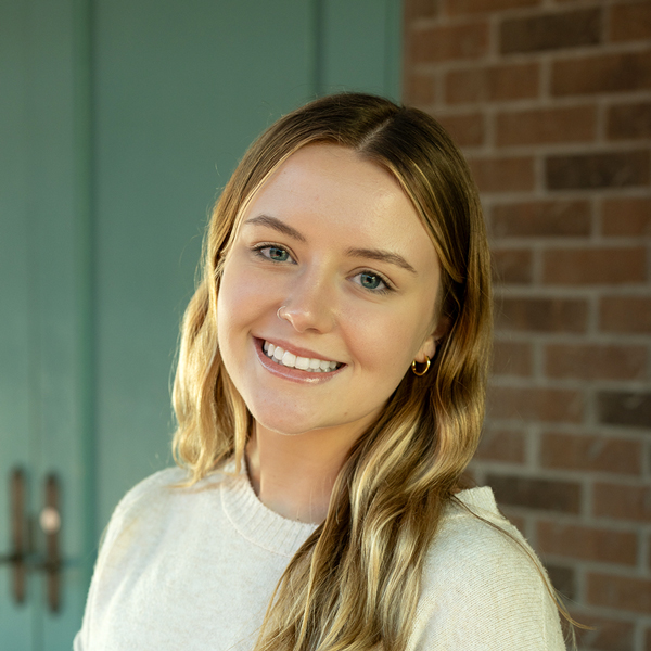 Olivia Schwendeman, Senior Living Director at Vitalia Mentor, smiling in a professional outdoor headshot with long blonde hair, gold hoop earrings, and softly blurred teal door and brick details in the background.