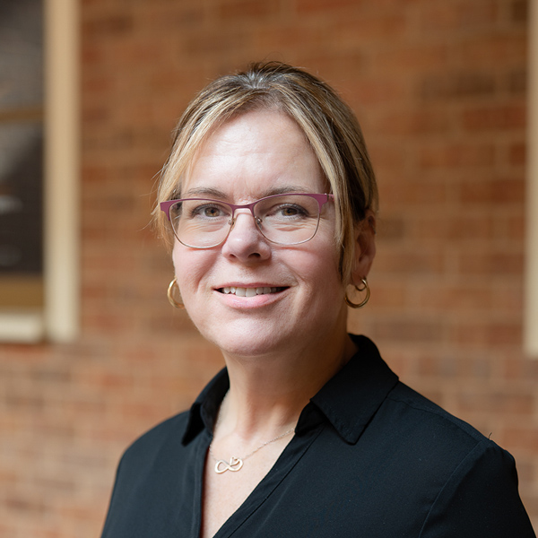 Kimberly “Kim” Semple, Wellness Director at Vitalia Mentor, looking at the camera in a professional outdoor headshot with blonde hair pulled back, pink glasses, gold hoop earrings, a black blouse, and a softly blurred brick background.