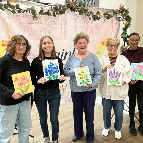 Residents and team members at Vitalia Mentor smile together, holding paintings featuring flowers.