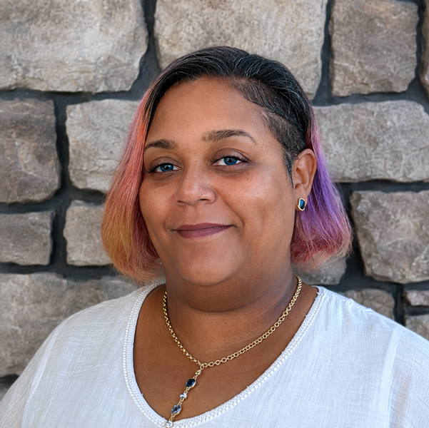 Tiffany Bobo, Memory Care Director at Vitalia Mentor, smiling in a professional headshot with short pink hair, a white blouse, and a stone wall background.